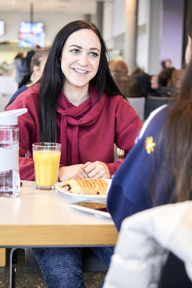 Brünette Studierende mit rotem Pullover sitzt in der Cafeteria der FH Bielefeld einer anderen Studierenden gegenüber und unterhält sich