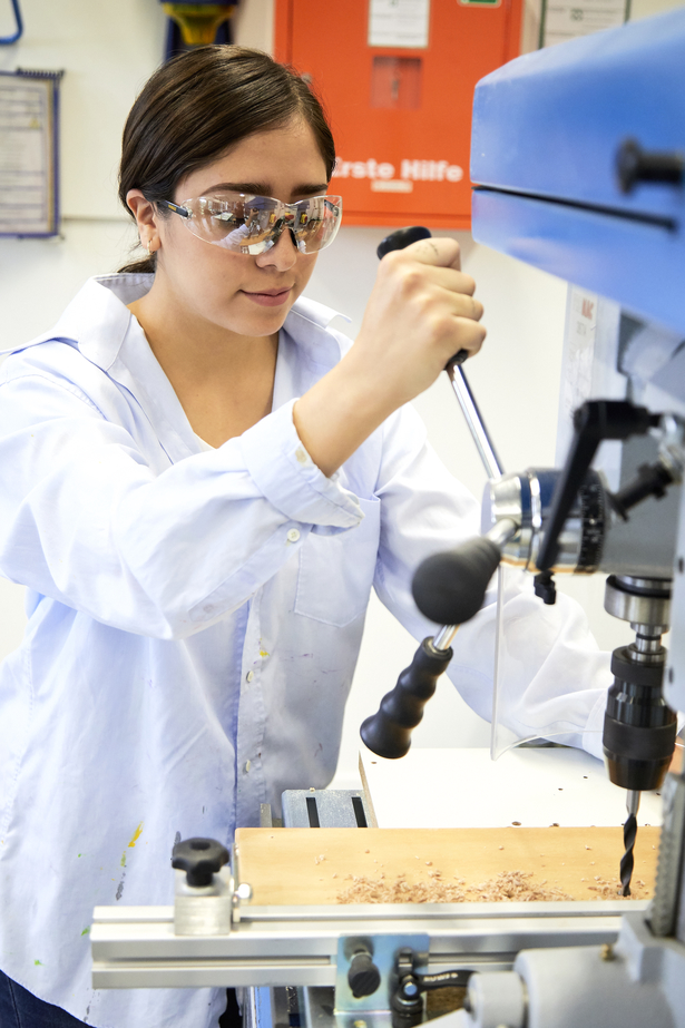 Brünette Studierende mit Kittel und Schutzbrille in einer Werkstatt an einem Gerät