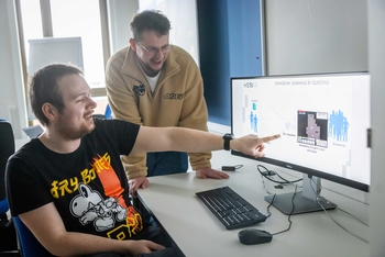 Ein Mann zeigt auf einem großen Computerbildschirm das Spiel neben ihm steht ein anderer Mann und schaut zu