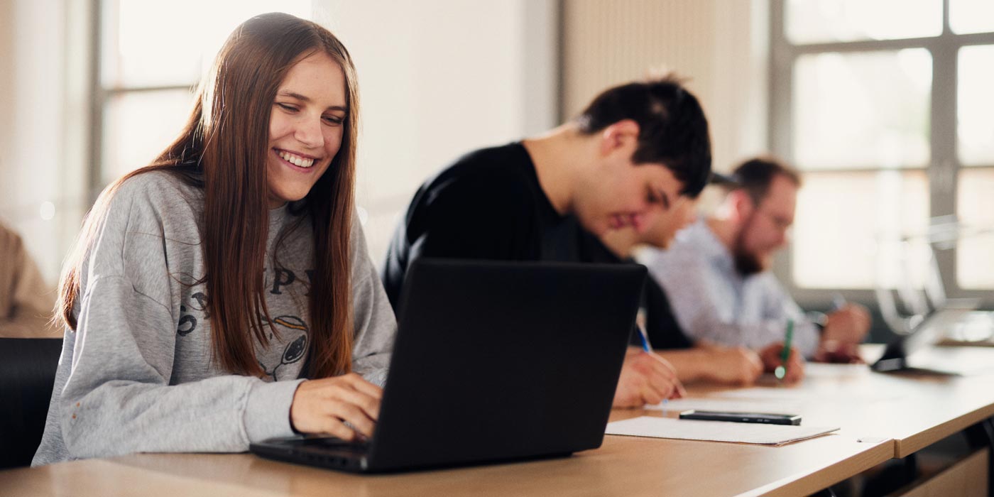 Eine Studentin sitzt an ihrem Laptop in einem Seminar und lächelt neben ihr sitzen weitere Studierende